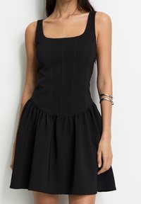 Robe noire avec encolure carrée, corsage ajusté avec des coutures verticales et jupe évasée avec tissu froncé pour plus de volume.