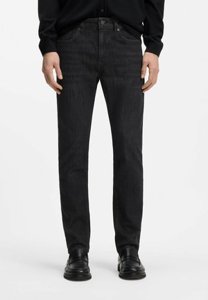 Vue de face d'une personne portant un jean noir à jambes droites, des mocassins noirs et une chemise noire à manches longues, se tenant devant un fond blanc.