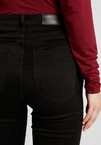 Zwarte jeans met een gladde textuur, voorzien van een hoge tailleband, twee achterzakken en gestikte detailing. Het bovenstuk is een rood langärmige shirt.
