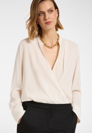 Blouse en beige clair, fabriquée en tissu doux, avec un col en V et des manches longues, assortie à un pantalon noir et un collier texturé doré.