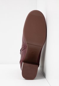 Zapato de cuero burdeos con suela de goma texturizada, que presenta un tacón cuadrado y un logo de marca en relieve. El zapato tiene un acabado suave y un diseño estilizado.