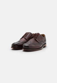Chaussures brogue en cuir marron avec un design à bouts ailés, des perforations décoratives et une finition brillante, accompagnées d'un talon empilé. Fermeture à lacets sécurisée.