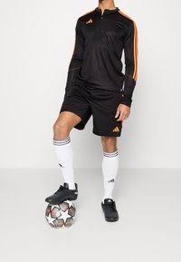 Person i svart och orange fotbollsdräkt som står med ena foten på en vit, svart och orange fotboll mot en neutral bakgrund.