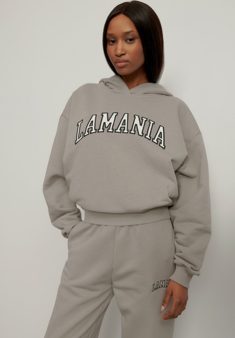 Hanorac gri cu glugă pe care scrie "LAMANIA" în litere negre, asociat cu pantaloni de trening gri asortați. Material neted, croială casual, manșete cu riburi.