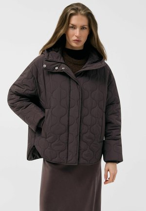 Kobieta ubrana w ciemnobrązową pikowaną kurtkę na zatrzaski oraz pasującą spódnicę, stojąca na tle jednolitego jasnego tła.