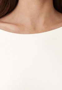 Ribbad, off-white topp med en bred halsringning. Tyget verkar mjukt med en diskret textur och visar en åtsittande design.