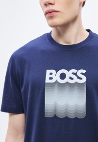 Temno modra bombažna majica z napisom "BOSS" v krepki beli pisavi in sivim valovitim prelivom pod besedilom.