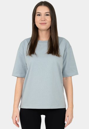 Woman with long brown hair wearing light gray short-sleeve T-shirt and black pants, standing against plain light background.