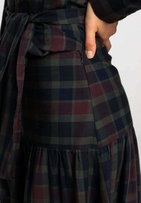 Robe à carreaux sombres présentant des teintes de bleu marine, de bordeaux et de vert. Détails notables incluant une taille nouée et une fermeture éclair sur le côté. Tissu texturé avec un ourlet évasé.
