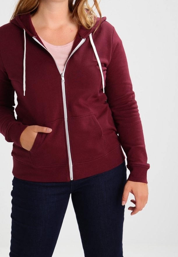 Femme portant un sweat à capuche bordeaux zippé avec cordons blancs et jean bleu marine, main dans la poche, sur un fond clair uni.