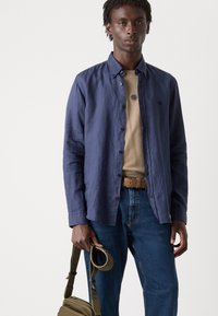 Marineblå skjorte med strukturelt stoff, beige t-skjorte under, blå jeans, brun belte og oliven grønn veske med justerbar stropp.