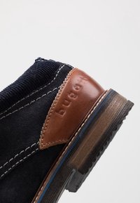 Botte en suède noir avec un contrefort en cuir marron, couture blanche et dessus texturé avec des détails nervurés. Semelle semblable à du bois avec une adhérence en caoutchouc.