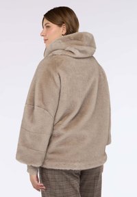 Beżowa kurtka z imitacji futra z wysokim kołnierzem i luźnym krojem. Długie, szerokie rękawy i miękka faktura. Brak widocznych zapięć.