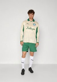 Puma långärmad tröja i krämfärg med gröna detaljer, har "KidSuper"-logotyp; kombinerad med gröna shorts, vita strumpor och svarta fotbollsskor.