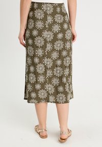 Jupe midi vert olive avec des motifs floraux blancs, en tissu doux, avec une fente latérale et une coupe décontractée. Associée à des sandales beiges.
