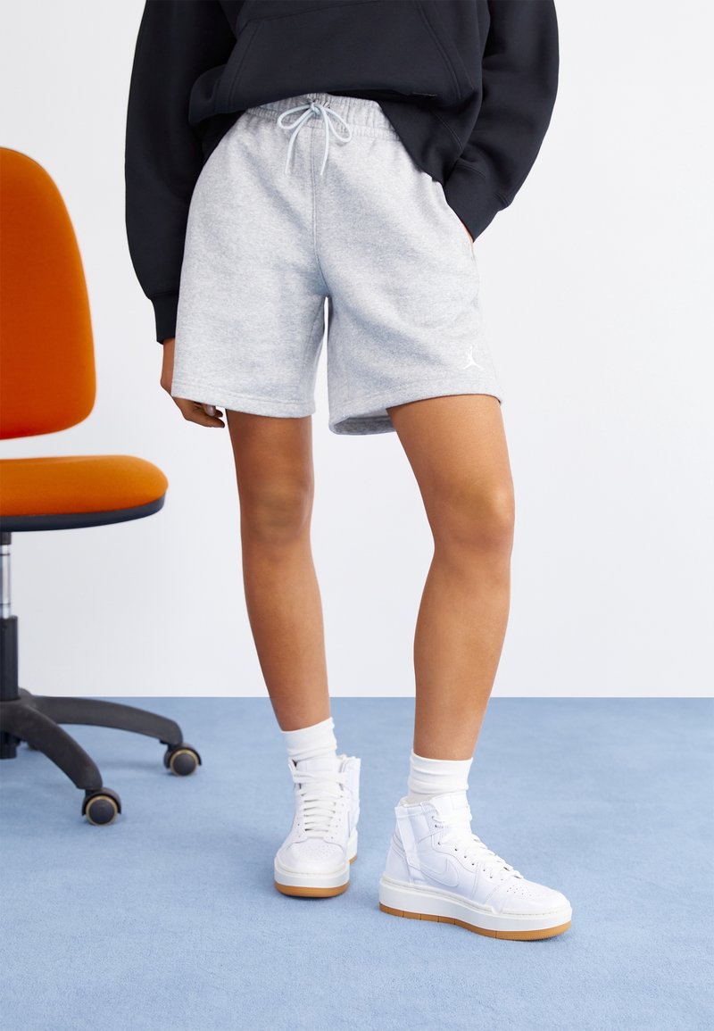 Personne portant un short gris clair à cordon, un sweat-shirt noir, des baskets blanches montantes et des chaussettes blanches, debout sur un tapis bleu près d'une chaise de bureau orange.