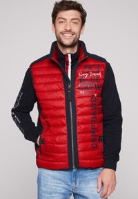 Gilet trapuntato rosso con maniche nere, chiusura frontale con zip, tasche laterali e dettagli testuali. Presenta un colletto alto e texture a contrasto.
