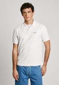 White polo shirt with a textured pattern of small blue anchors, featuring a collar and short sleeves. Logo on the chest. Casual fit.