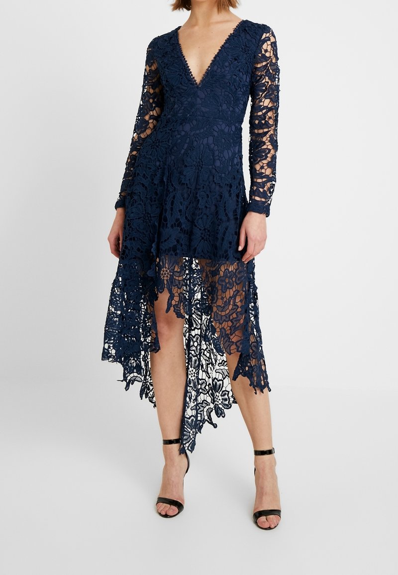 Femme portant une robe en dentelle bleu marine à manches longues, col en V profond, ourlet asymétrique, et des talons noirs à bride à la cheville, debout sur fond blanc.