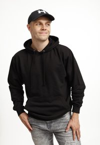 Giovane uomo che indossa un hoodie nero e un cappello, in piedi con le mani sui fianchi, che guarda leggermente di lato, sorridendo in modo sottile.