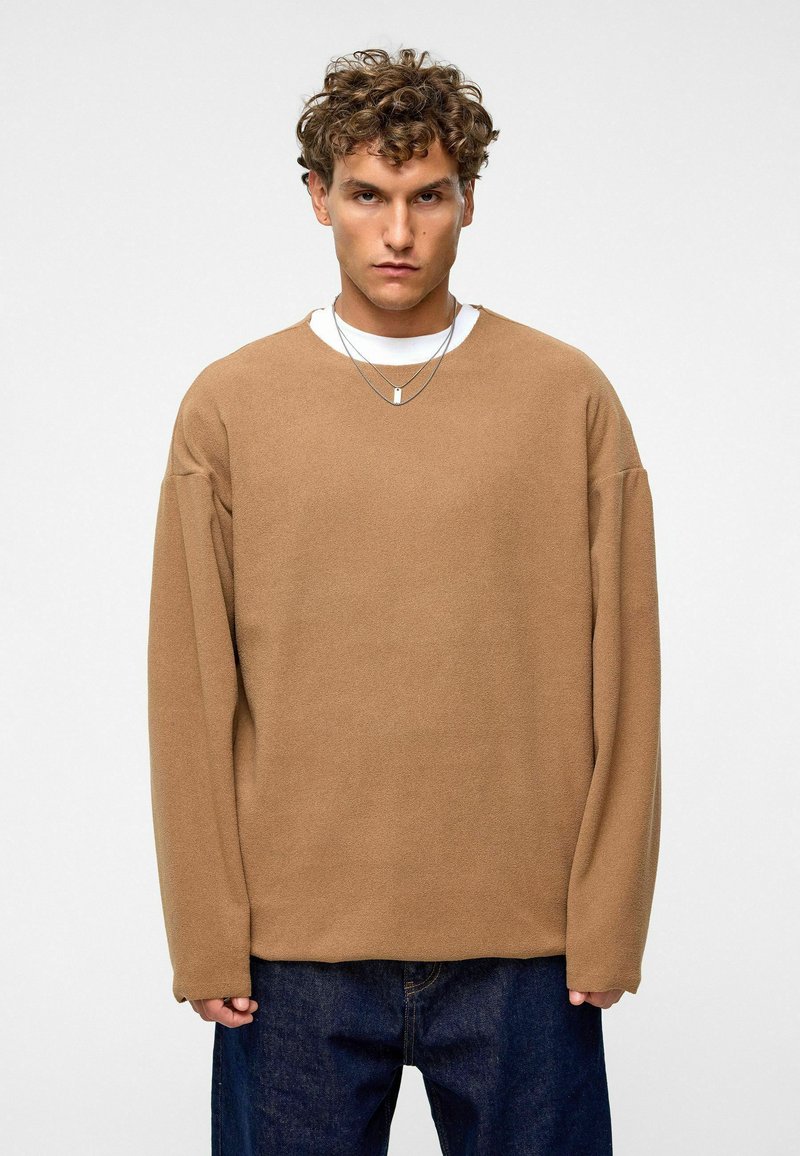 Bruine oversized longsleeve sweatshirt met een textuur, uitgerust met verlaagde schouders en een ronde halslijn, gelaagd over een wit shirt.