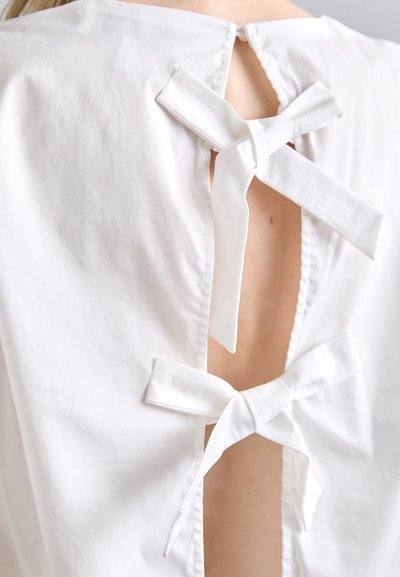 Fehér blúz hátsó nyílással, amelyet három szövetmasni rögzít. Simaság jellemzi, letisztult, minimalista dizájn és enyhén ívelt nyakkivágás.