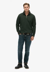 Sudadera de cremallera en verde oscuro con un cuello de cuadros, combinada con jeans de mezclilla azul y botas negras de cordones. El modelo está de pie con las manos en los bolsillos.