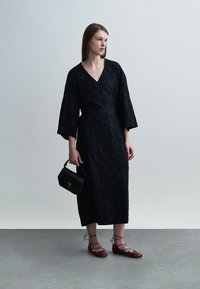 Robe portefeuille noire texturée avec manches longues, longueur mi-mollet. Portée avec des ballerines bordeaux à lanières et un petit sac à main noir.