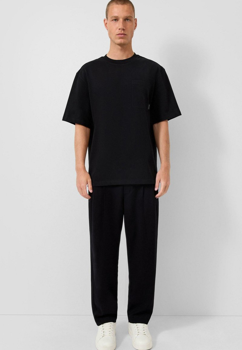 Mladý muž stojí opřený o šedou zeď, má na sobě velké černé tričko s kapsou, volné černé kalhoty a bílé tenisky.