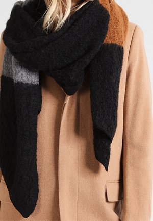 Femme portant une écharpe épaisse et texturée à blocs de couleurs noir, gris et marron sur un manteau en laine couleur chameau.