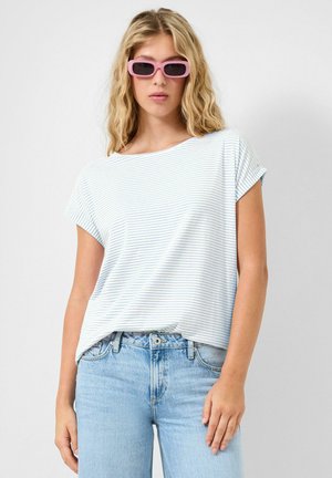Femme aux cheveux blonds ondulés portant des lunettes de soleil roses, un t-shirt blanc à rayures bleu clair et un jean bleu clair sur un fond uni.
