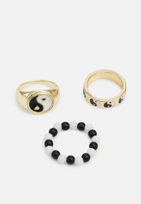 Três anéis apresentam: um anel de ouro com um design yin-yang em preto e branco, uma banda lisa de ouro e um anel de contas em preto e branco.