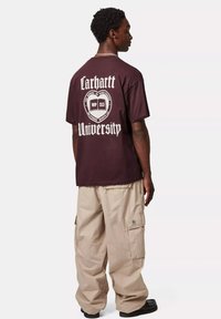 Camiseta de algodón color marrón con texto gráfico blanco que dice "Carhartt University" en la parte de atrás, combinada con pantalones cargo holgados color beige.
