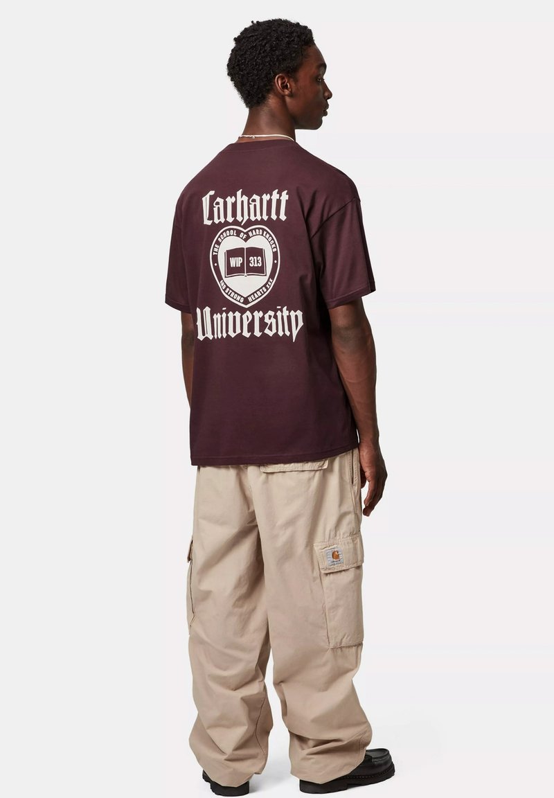 T-shirt di cotone bordeaux con testo grafico bianco che legge "Carhartt University" sul retro, abbinato a pantaloni cargo beige larghi.