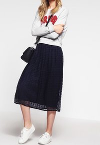 Sudadera gris con texto rojo, combinada con una falda midi plisada azul marino y zapatillas blancas. Bolso bandolera negro. Conjunto sencillo y casual.