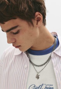Gestreiftes, leichtes Hemd über einem blauen T-Shirt, verziert mit geschichteten Silberketten und einem Anhänger. Nahaufnahme eines jungen männlichen Models.