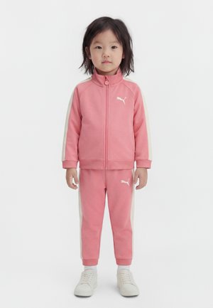 Young child standing, wearing a pink Puma tracksuit with white stripes down the sleeves and legs, and white sneakers.
