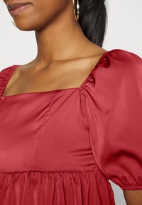 Vestido de cetim vermelho com decote quadrado, mangas bufantes e detalhes franzidos. Textura suave e caimento elegante. Brincos de argola dourados visíveis.