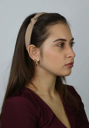 Bandeau beige en tissu avec un détail noué, porté par une personne aux longs cheveux bruns, sur un fond clair uni. Boucles d'oreilles créoles en or.