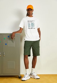 Witte t-shirt met groene grafische tekst, groene cargoshorts, oranje pet, witte sokken en zwart-geaccentueerde witte sneakers. Metalen locker op de achtergrond.