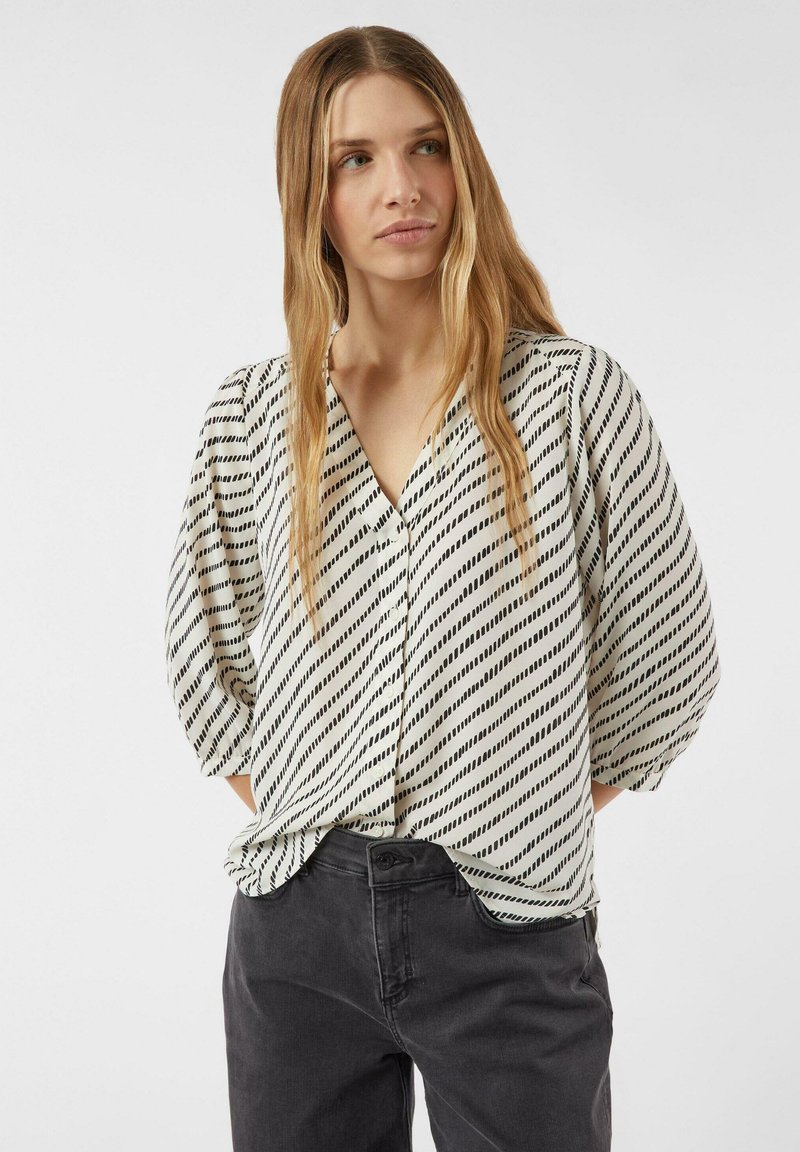 Blouse ample en tissu léger avec un fond blanc et des rayures diagonales noires. Encolure en V et manches courtes bouffantes.