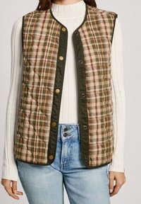 Vadderad rutig väst i jordnära toner, med grön kantning och tryckknappar. Lagd över en vit ribbad tröja. Blå jeans syns nedanför.