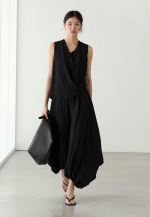 Frau, die drinnen geht und ein ärmelloses schwarzes, drapiertes Oberteil, weite schwarze Hosen, schwarze Sandalen trägt und eine große schwarze Handtasche hält.