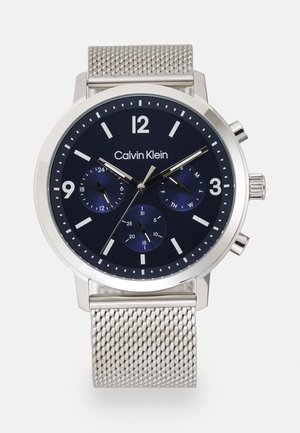 Calvin Klein silver metal mesh watch with black dial, three subdials for 24-hour, date, day, and silver hour markers and hands.