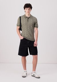 Zelena polo majica s uzorkom, crne kratke hlače i bijele tenisice s tamnim naglascima. Model stoji s rukama u džepovima, s torbom preko ramena.
