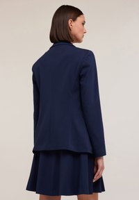 Marineblauwe blazer met een getailleerde pasvorm, voorzien van lange mouwen en lichte schoudervullingen. Gelaagd over een geplooide marineblauwe rok.