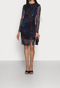 Zwarte doorzichtige jurk met lange mouwen, versierd met een gekleurd bloemenpatroon, geraffineerde details, een zijsplit, en gecombineerd met zwarte open teen-hakken.