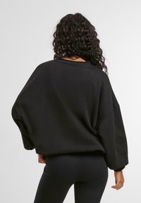 Svart oversized genser med vid hals, lange ermer, myk tekstur og et subtilt brodert design på venstre erme.