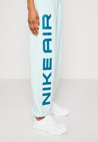 Světlé modré tepláky s velkým tmavě modrým logem "NIKE AIR" běžícím vertikálně po levé noze, doplněné bílými sportovními teniskami.