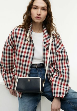 Jonge vrouw draagt een rood-wit-blauw geruit jasje, wit T-shirt, blauwe spijkerbroek en een zwart-witte crossbodytas met gouden logo.